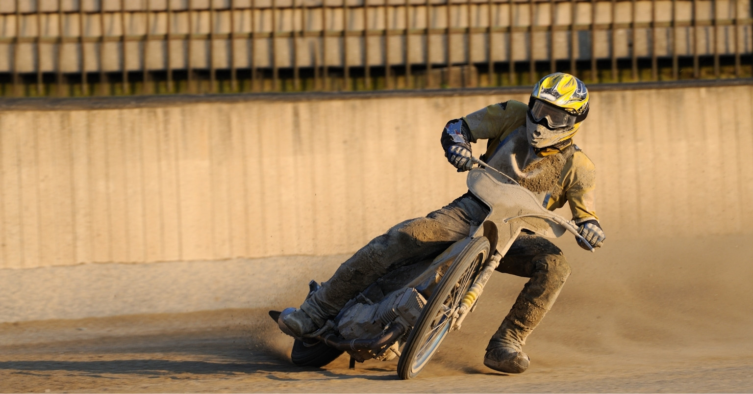 A speedway rider in a yellow helmet leans into a corner on a dirt track